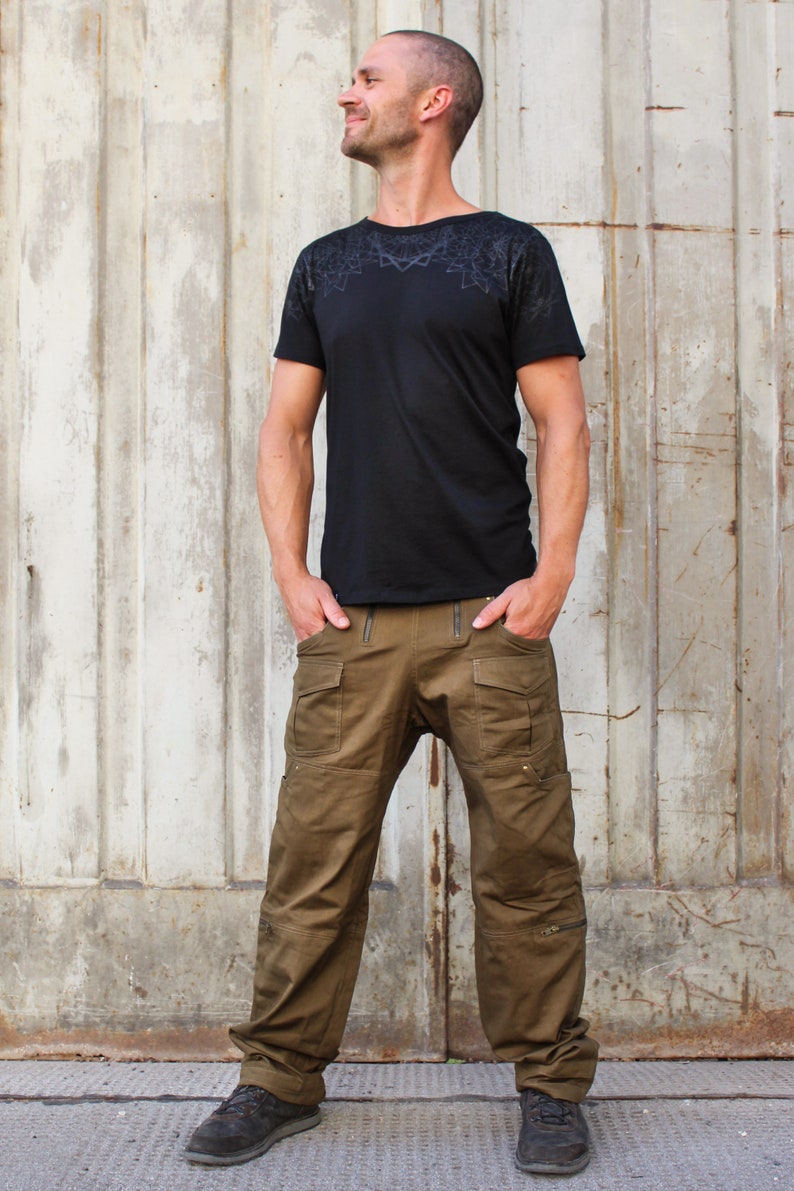 May include: A man wearing a black t-shirt with a geometric pattern and brown cargo pants. He is standing in front of a metal wall.
