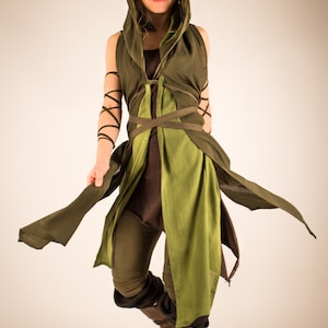 May include: A woman wearing a green hooded tunic with brown pants and brown boots. The tunic has a green and brown color scheme and is tied with a brown belt.