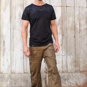May include: A man wearing a black t-shirt with a geometric pattern and brown cargo pants. He is standing in front of a white wooden wall.