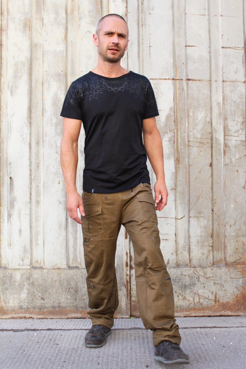 May include: A man wearing a black t-shirt with a geometric pattern and brown cargo pants. He is standing in front of a white wooden wall.
