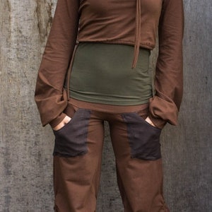 May include: A woman wearing a brown cowl neck top with a green undershirt and brown pants with black pockets. She is standing in front of a wooden wall.