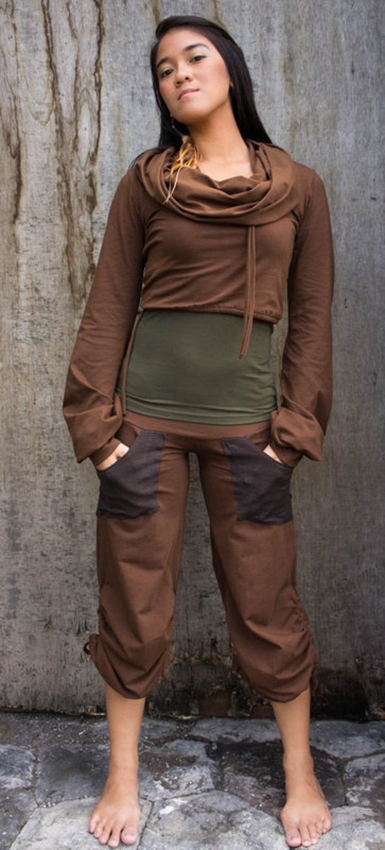 May include: A woman wearing a brown cowl neck top with a green undershirt and brown pants with black pockets. She is standing in front of a wooden wall.
