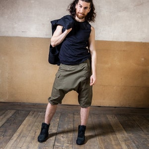 May include: A man wearing a black tank top, green cargo shorts, and black sneakers. He is holding a black jacket over his shoulder. He has long brown hair and a beard.