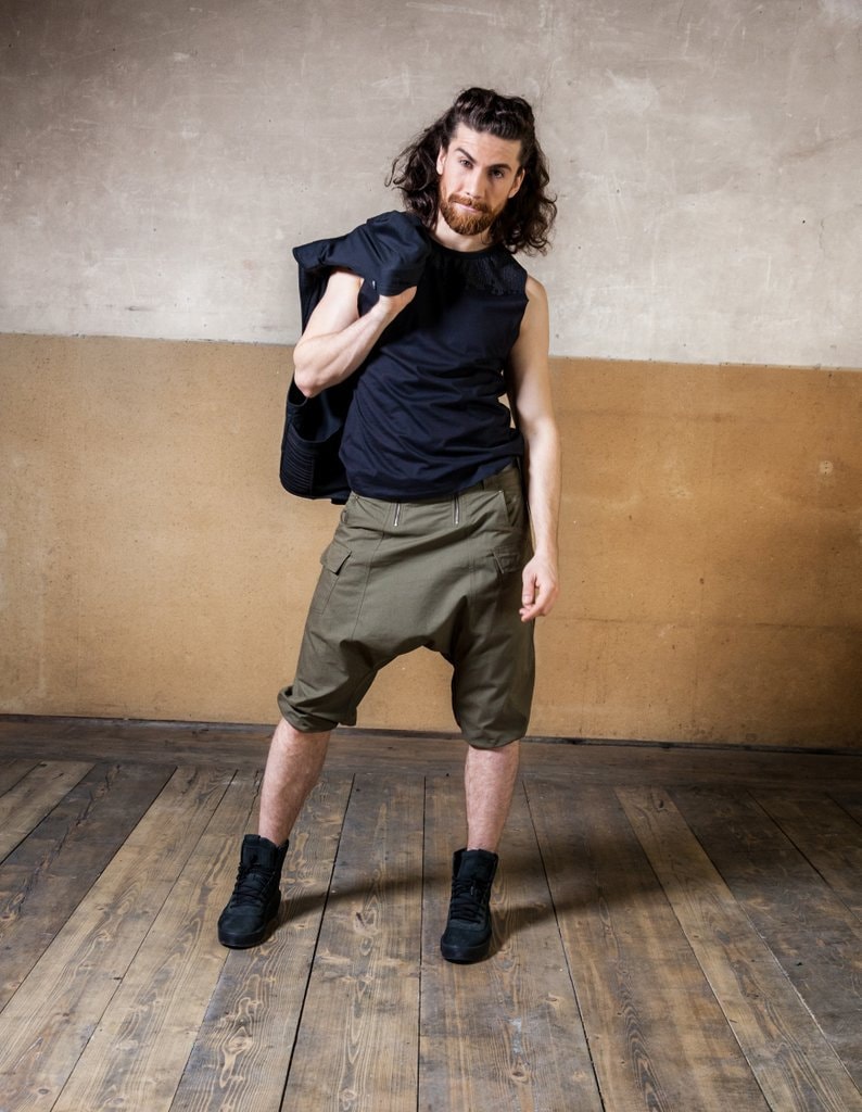 May include: A man wearing a black tank top, green cargo shorts, and black sneakers. He is holding a black jacket over his shoulder. He has long brown hair and a beard.
