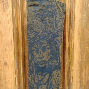 May include: A tall, olive green wine bottle bag with a detailed blue and gold illustration of a face with a headdress. The bag has a matching olive green strap and is made of a textured fabric. The bag is hanging in front of a wooden door.