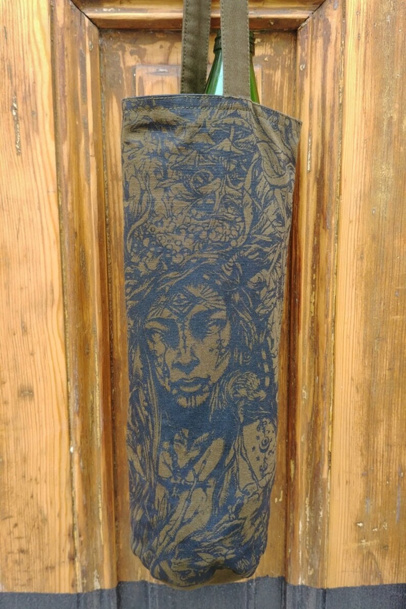May include: A tall, olive green wine bottle bag with a detailed blue and gold illustration of a face with a headdress. The bag has a matching olive green strap and is made of a textured fabric. The bag is hanging in front of a wooden door.