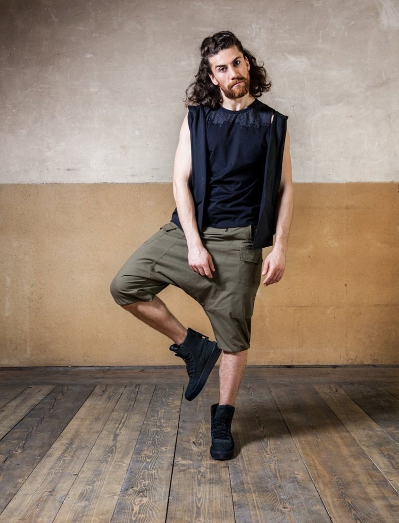May include: A man wearing a black sleeveless shirt and green cargo shorts stands with one leg raised. He is wearing black boots and has long brown hair.