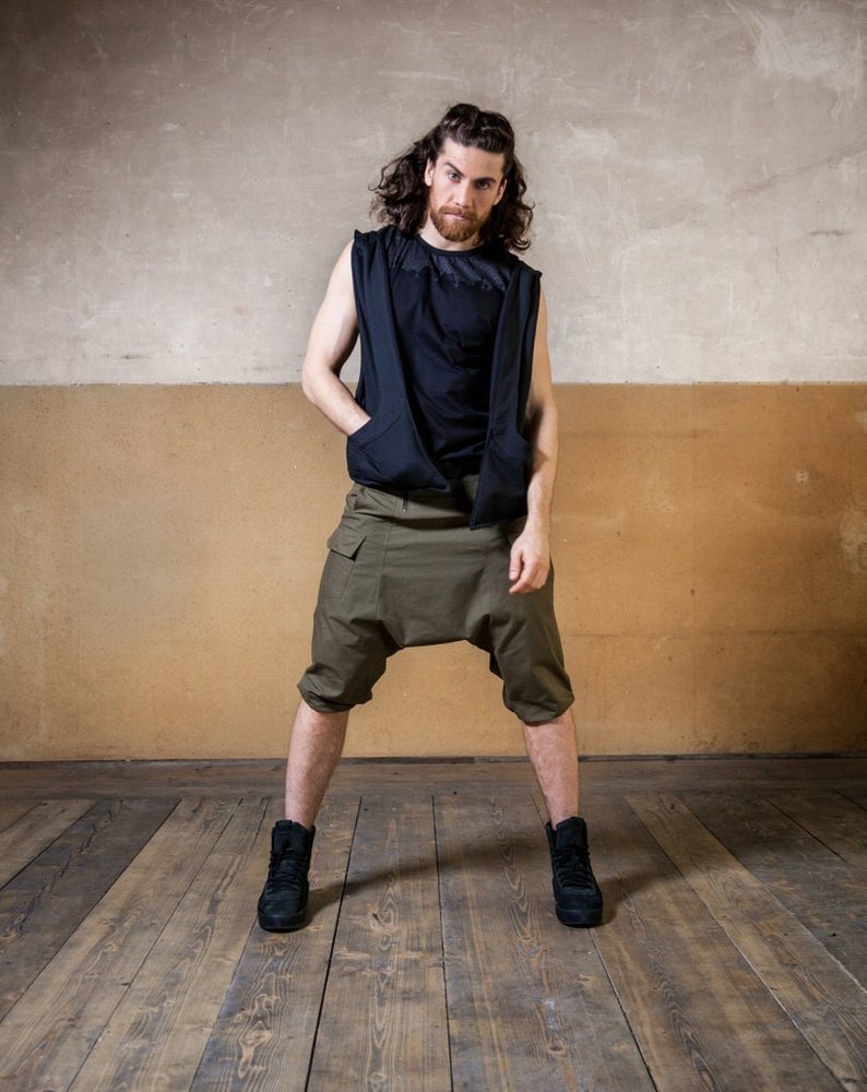 May include: A man wearing a black sleeveless shirt with a zipper and a pair of olive green cargo shorts. He is standing on a wooden floor.