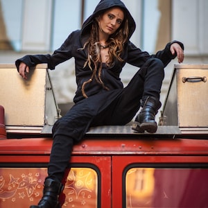 May include: A woman with long brown dreadlocks wears a black hooded jacket and black pants. She sits on the roof of a red vehicle with a metal box behind her.