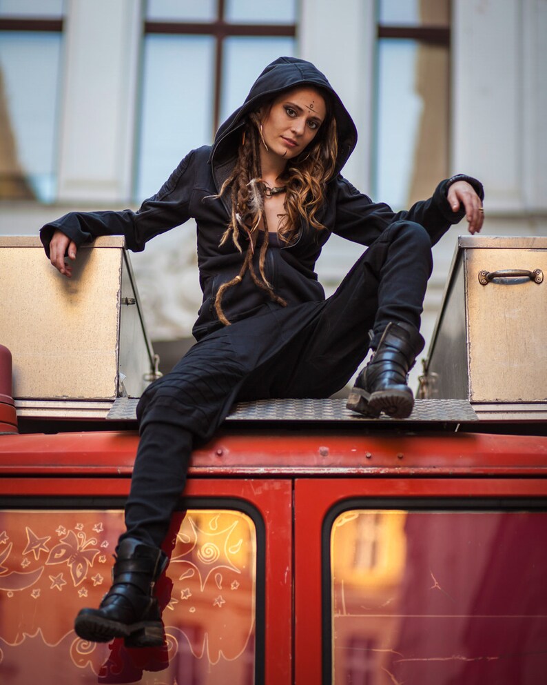 May include: A woman with long brown dreadlocks wears a black hooded jacket and black pants. She sits on the roof of a red vehicle with a metal box behind her.
