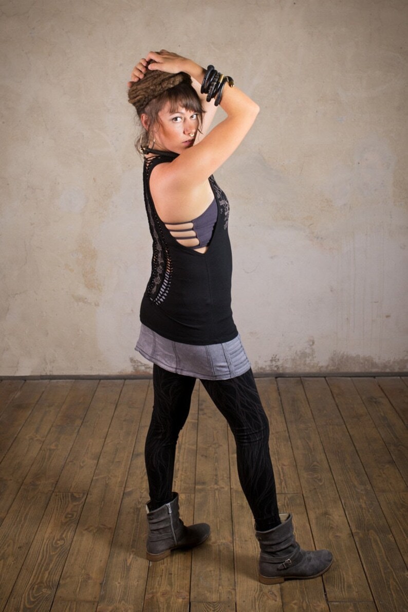 May include: A woman wearing a black tank top with cut-out detailing, a gray skirt, and black leggings with a pattern. She is also wearing gray boots and multiple black bracelets. Her hair is styled in dreadlocks.