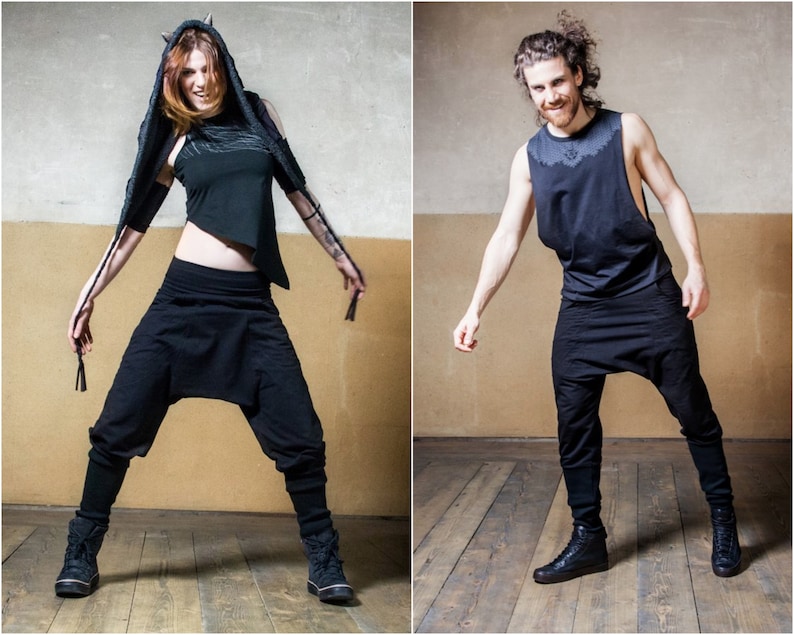 Puede incluir: Dos personas con ropa negra y zapatos negros posan en un estudio. La mujer lleva una camiseta negra con capucha, una bufanda larga y pantalones negros. El hombre lleva una camiseta negra sin mangas y pantalones negros.