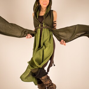 May include: A woman wearing a green hooded tunic with brown straps and a brown undershirt. She is holding the edges of the tunic, which is flowing in the air. She is wearing brown boots.