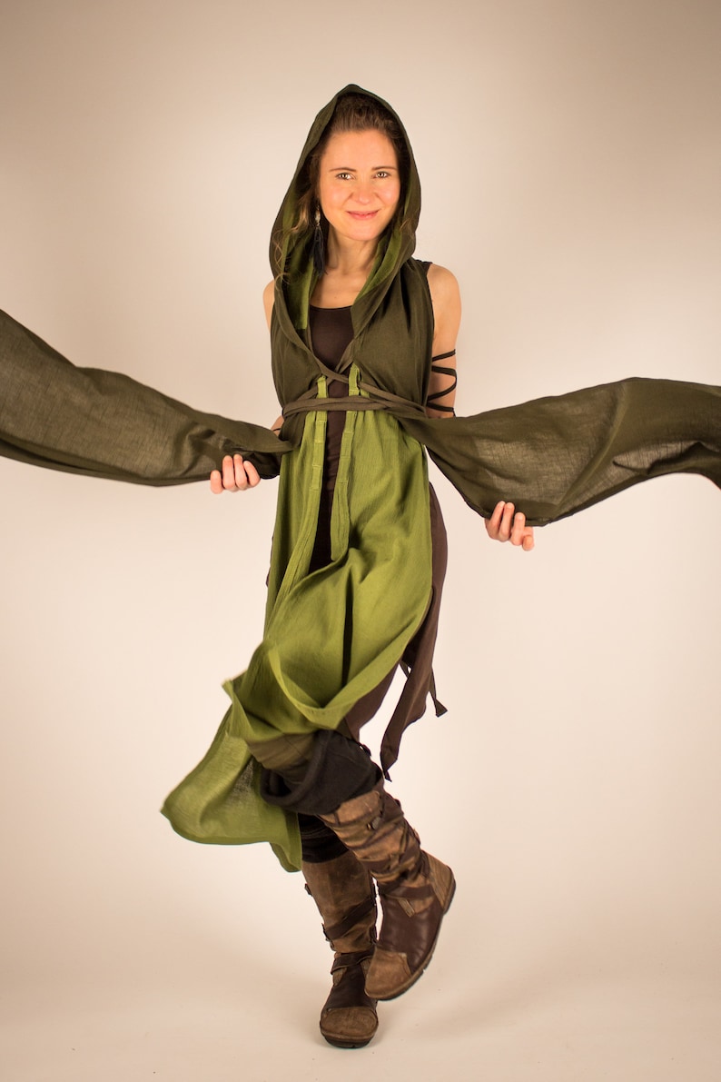 May include: A woman wearing a green hooded tunic with brown straps and a brown undershirt. She is holding the edges of the tunic, which is flowing in the air. She is wearing brown boots.