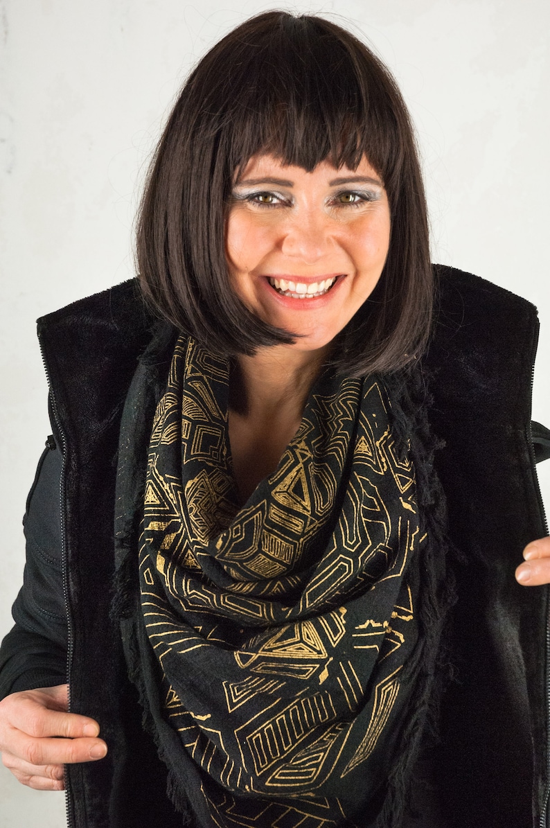 May include: A woman with dark brown hair wearing a black jacket with a gold and black geometric patterned scarf.