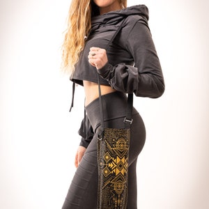 May include: Black water bottle carrier with a geometric gold design. The carrier is held by a black strap. The carrier is worn over the shoulder. The person in the image is wearing a dark gray cropped hoodie and leggings.