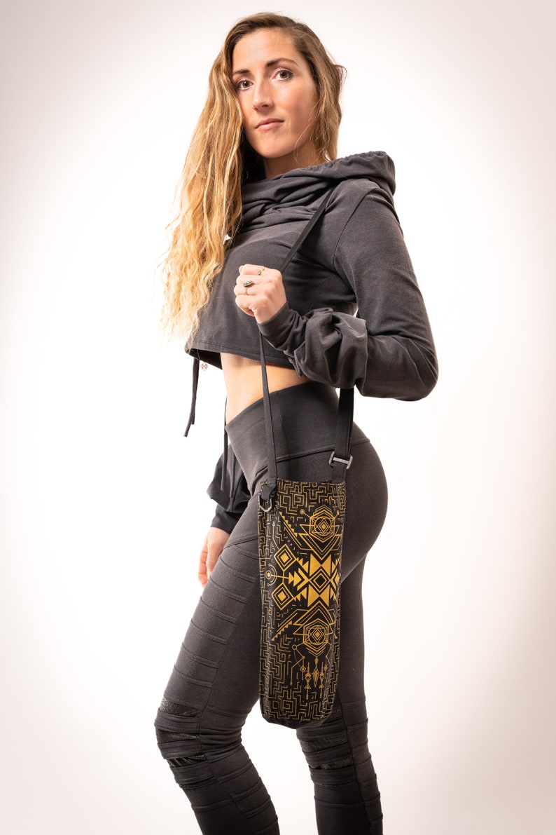 May include: Black water bottle carrier with a geometric gold design. The carrier is held by a black strap. The carrier is worn over the shoulder. The person in the image is wearing a dark gray cropped hoodie and leggings.