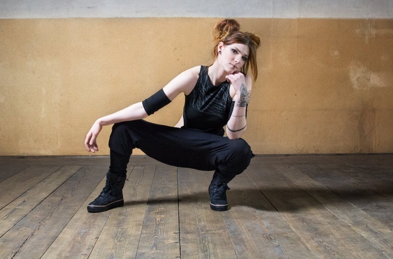 Puede incluir: Una mujer con pantalones negros y un top negro sin mangas con un dise&ntilde;o texturizado. Est&aacute; sentada en un suelo de madera con los brazos cruzados y mirando a la c&aacute;mara.