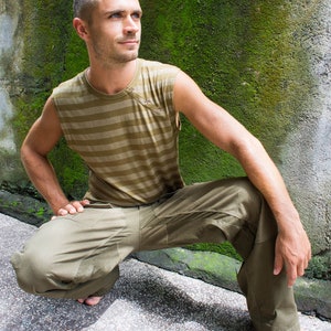 May include: A man wearing a striped sleeveless shirt and khaki cargo pants is squatting in front of a mossy wall.