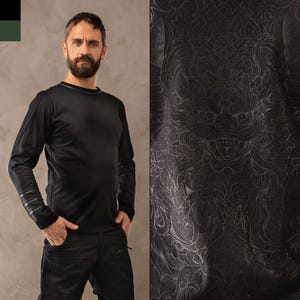 May include: Black long-sleeved shirt with a subtle silver graphic of a mythical creature on the front. The graphic is detailed and intricate, with a fierce expression.