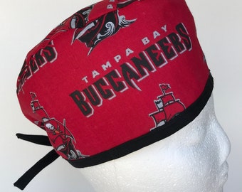 May include: Red and black surgical scrub hat with a repeating pattern of the Tampa Bay Buccaneers logo. The logo features a pirate ship, skull and crossbones, and the words "Tampa Bay Buccaneers".