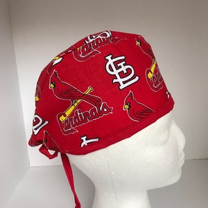 May include: Red surgical scrub hat with a white St. Louis Cardinals logo and a red cardinal bird with a baseball bat. The hat has two long ties.