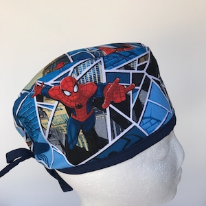 May include: Blue and red surgical scrub hat with a cartoon image of Spiderman in a comic book style design. The hat has blue ties.