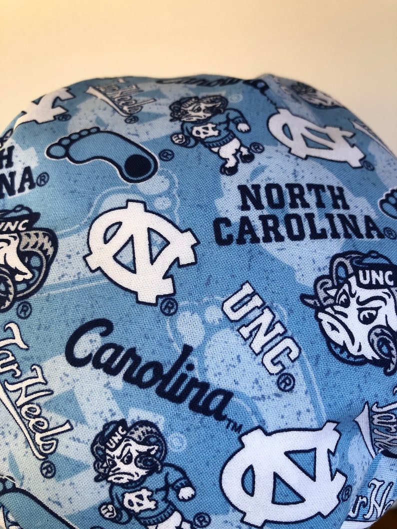 May include: Blue and white fabric with a repeated pattern of the University of North Carolina Tar Heels logo, the words "North Carolina" and "UNC", and a cartoon ram mascot. The fabric also features a blue footprint.