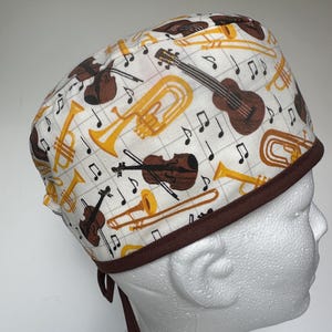 Può includere: Un cappello chirurgico bianco con un motivo ripetuto di strumenti musicali, tra cui violini, trombe e tromboni, con note musicali. Il cappello ha un bordo e lacci marroni.