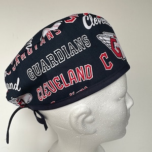 May include: A blue and red surgical scrub hat with the Cleveland Guardians logo and the text "Cleveland Guardians" and "C" printed on it.