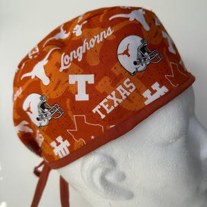 Op de afbeelding: Een oranje en witte scrub-muts met een Texas Longhorns-thema. De muts heeft een herhalend patroon van het Texas Longhorns-logo, de woorden "Longhorns" en "Texas", en een voetbalhelm met een Longhorn-stier.