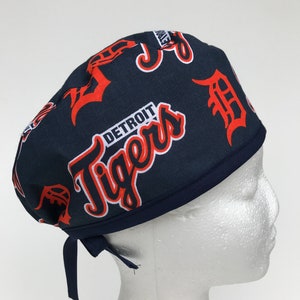 May include: A navy blue surgical scrub hat with a white and orange Detroit Tigers logo print. The hat has two ties to adjust the fit.
