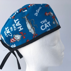May include: Blue scrub hat with a black trim and black ties. The hat features a repeating pattern of the Cat in the Hat character from Dr. Seuss books. The text "The Cat in the Hat" is printed on the hat.