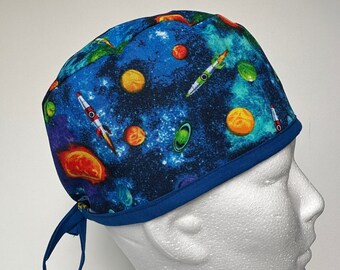 Könnte beinhalten: Eine blaue OP-Mütze mit einem Weltraum-Motiv mit Planeten, Sternen und Raketen. Die Mütze hat einen blauen Saum und eine Bindekordel.