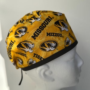 May include: A yellow and black surgical scrub hat with a repeating pattern of the University of Missouri logo, tiger mascot, and the words "Missouri", "Mizzou", and "Tiger".