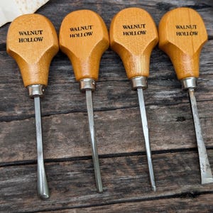 May include: Four wood carving tools with light brown wooden handles, each stamped with the text WALNUT HOLLOW. The tools feature silver metal ferrules and distinct steel blade shapes, including a gouge, a V-tool, and a flat chisel tip.