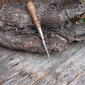May include: A vintage wood chisel with a wooden handle and a metal blade. The chisel is resting on a weathered wooden surface and a piece of textured bark.