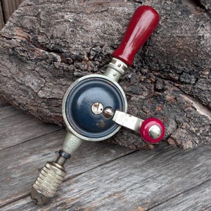 May include: Vintage hand drill with a dark blue body, silver metal components, and a red wooden handle. The drill bit is silver and textured. The handle and the side crank have red accents. The tool is set against a weathered wood background.