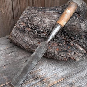 May include: A vintage wood chisel with a metal blade and a wooden handle. The chisel is resting on a weathered wooden surface, with a piece of dark, textured wood in the background. The tool appears well-used.