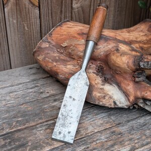 May include: A wood chisel featuring a flat, rectangular metal blade with visible signs of wear and oxidation, connected to a cylindrical wooden handle with a dark metal ferrule at the base of the grip, showing a natural wood grain texture.