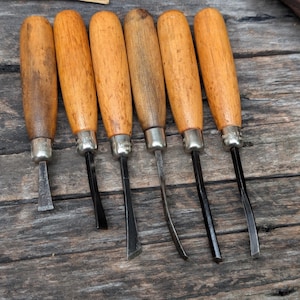 May include: A set of six wood carving tools featuring light brown wooden handles and silver-toned metal ferrules. Each tool has a distinctively shaped steel blade, including straight, angled, and curved profiles for detailed woodworking tasks.