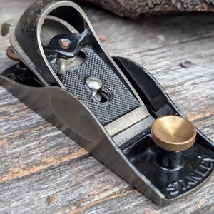 May include: A vintage Stanley block plane, featuring a black body with a textured grip area and a brass adjustment knob. The blade is silver, and the tool is set on a wooden surface.