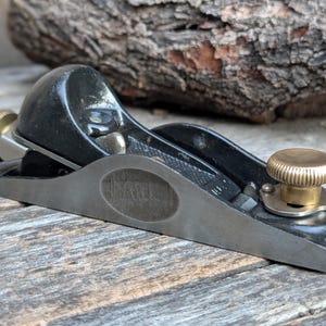 May include: A small, black hand plane with a brass knob and adjustment wheel. The plane has a silver-colored sole and a curved body. The word "MILL" is stamped on the side. The tool is designed for woodworking.