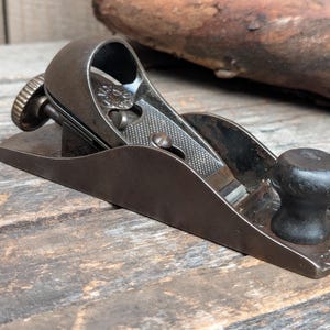May include: A vintage metal block plane featuring a dark brown finish, a rounded black knob at the front, and an adjustable screw mechanism at the rear. The tool has a textured metal lever cap and a flat base, showing signs of age and wear.