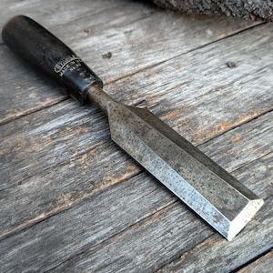 May include: A vintage wood chisel with a black handle and a metal blade. The chisel is lying on a weathered wooden surface. The blade is angled and has a sharp edge.