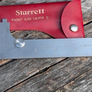 May include: A stainless steel Starrett pocket slide caliper, 5 inches long, with a measurement scale. The caliper is next to a red leather case with the Starrett logo and product information printed in gold lettering.