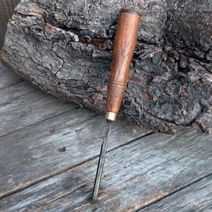 May include: A woodworking chisel with a wooden handle and a metal blade. The handle is a warm brown color with a brass ferrule. The blade is a silver color. The chisel is resting on a weathered wooden surface.