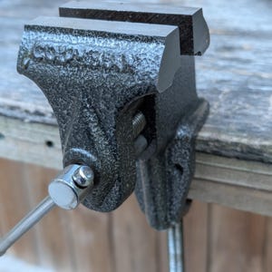 May include: A heavy-duty, black metal vise with a textured finish. The vise features a handle for adjusting the jaws, which are open in the image. The vise is designed to clamp and hold objects securely for various tasks. It is mounted on a wooden surface.