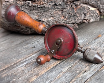 Puede incluir: Un taladro manual vintage con un cuerpo de metal rojo y un mango de madera. El taladro tiene una manivela para operar y un portabrocas de metal para sujetar las brocas. El mango es de color marrón claro y el taladro está sobre una superficie de madera.