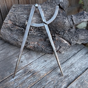 May include: A vintage metal compass, featuring two pointed legs and a curved arm, rests on a weathered wooden surface. The compass is silver-toned with signs of age, suggesting it's a tool for measuring or drawing circles. The background includes a piece of dark, textured wood.